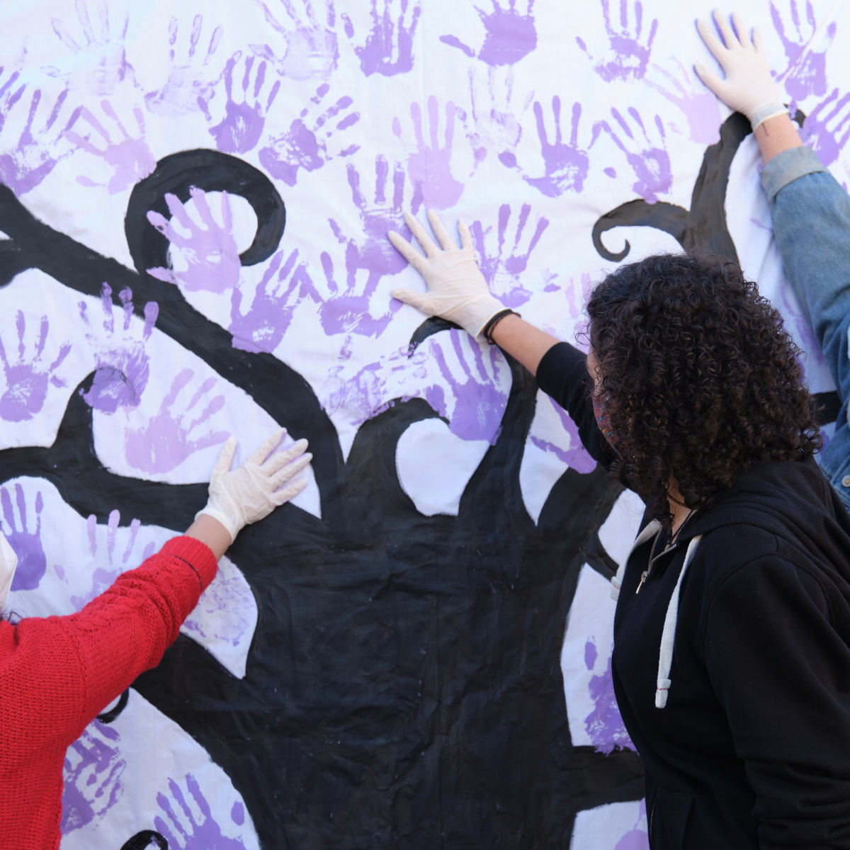 Tres mujeres participan en un mural pintando manos violetas por el Día Internacional de la Mujer